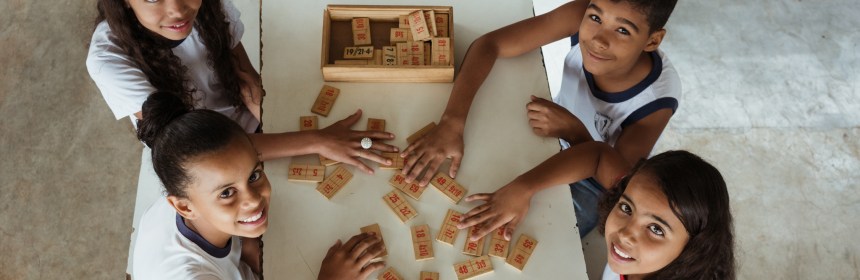 A imagem mostra cinco crianças sentadas ao redor de uma mesa branca, sorrindo e olhando para cima, em direção à câmera. Elas estão brincando com peças de madeira que contêm operações matemáticas e números. Sobre a mesa, também há três cadernos coloridos com o título "Língua Portuguesa". O ambiente parece ser uma sala ou área escolar com piso de concreto. A cena transmite um clima de aprendizado e colaboração.